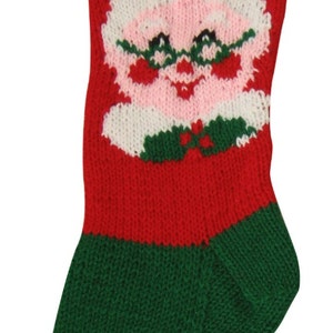 May include: A knitted Christmas stocking in red, green, and white. The top section features the letters "AKJ" in red on a white striped background. The stocking has a Santa Claus face design.