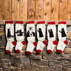 Op de afbeelding: Zes kerstsokken met rode manchetten en hielen, witte lijven en zwart-groene geborduurde ontwerpen. De sokken hangen aan een stenen muur voor een houten achtergrond. Elke sok heeft een andere kerststalscène.