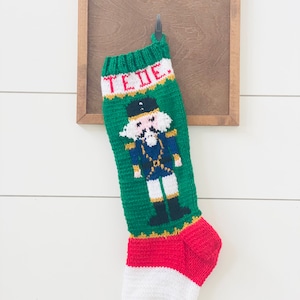 Puede incluir: Un calcetín navideño tejido en verde, rojo y blanco con un diseño de cascanueces. El calcetín está colgado de un gancho contra un marco de madera. El nombre "TEDIE" está tejido en la parte superior.