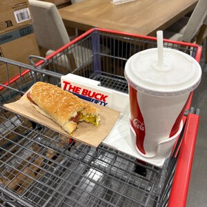 May include: A shopping cart with a hot dog on a paper tray and a Coca-Cola cup. The hot dog has a sesame seed bun, mustard, and relish. The cup is red and white with a straw. The cart has a sign that says "THE BUCK FIFTY".