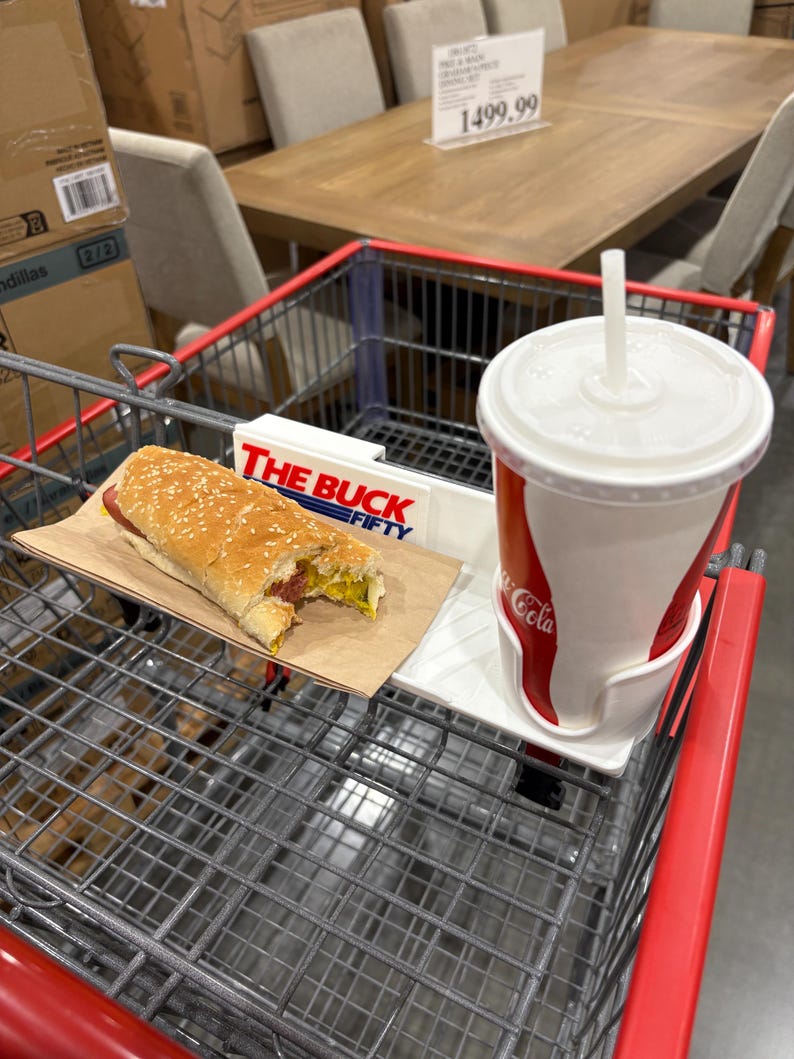May include: A shopping cart with a hot dog on a paper tray and a Coca-Cola cup. The hot dog has a sesame seed bun, mustard, and relish. The cup is red and white with a straw. The cart has a sign that says "THE BUCK FIFTY".