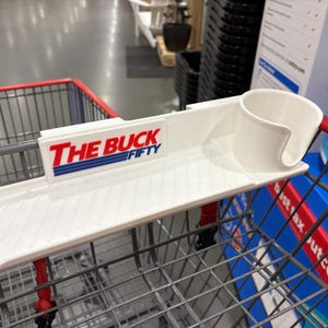 May include: A white plastic shopping cart accessory with a tray, cup holder, and the text "THE BUCK FIFTY" in red and blue. The accessory is designed to attach to a shopping cart.