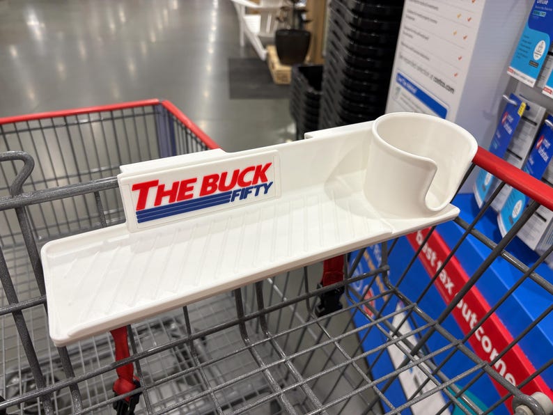 May include: A white plastic shopping cart accessory with a tray, cup holder, and the text "THE BUCK FIFTY" in red and blue. The accessory is designed to attach to a shopping cart.
