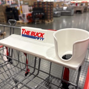 May include: A white shopping cart accessory with a cup holder and a small shelf. The text "THE BUCK FIFTY" is printed on the accessory in red and blue. The accessory is attached to a shopping cart.