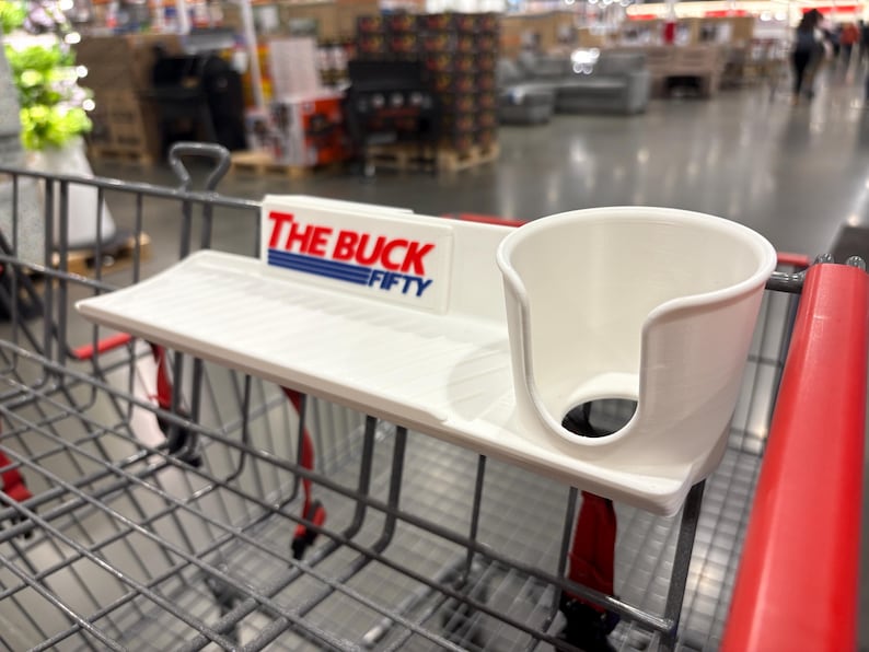 May include: A white shopping cart accessory with a cup holder and a small shelf. The text "THE BUCK FIFTY" is printed on the accessory in red and blue. The accessory is attached to a shopping cart.