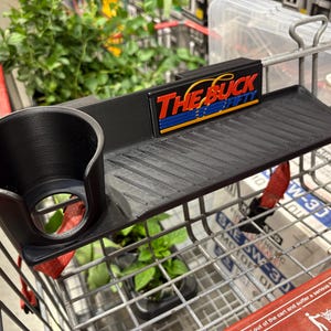 Puede incluir: Un accesorio negro para carrito de compras con un portavasos y una bandeja pequeña. El texto "THE BUCK FIFTY" se muestra en el accesorio. El accesorio está sujeto a un carrito.