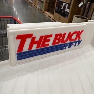 May include: White plastic shopping cart sign with the words "THE BUCK FIFTY" in red and blue lettering. The sign is attached to a white shopping cart.