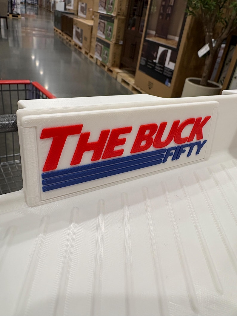 May include: White plastic shopping cart sign with the words "THE BUCK FIFTY" in red and blue lettering. The sign is attached to a white shopping cart.