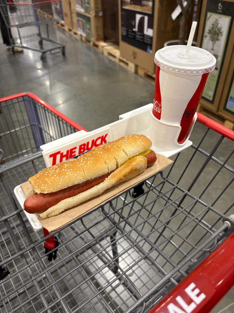 May include: A hot dog on a sesame seed bun with mustard, served on a paper tray, and a Coca-Cola drink with a straw, in a shopping cart. The cart has a white tray that says "THE BUCK".