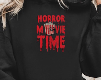 May include: Black hoodie with the text "HORROR MOVIE TIME" in red, featuring a popcorn bucket graphic. The text has a dripping blood effect. The hoodie has a hood and a front pocket.