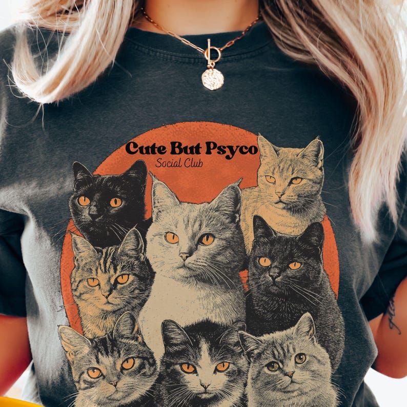 May include: A dark grey t-shirt featuring a graphic of a group of cats in a circle with a sunset background. The text "Cute But Psycho Social Club" is printed above the cats.