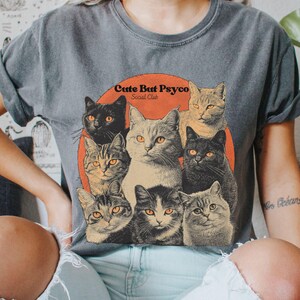 May include: A grey t-shirt with a vintage-style graphic of cats. The graphic features a group of cats with different coloured fur and the text "Cute But Psycho Social Club".