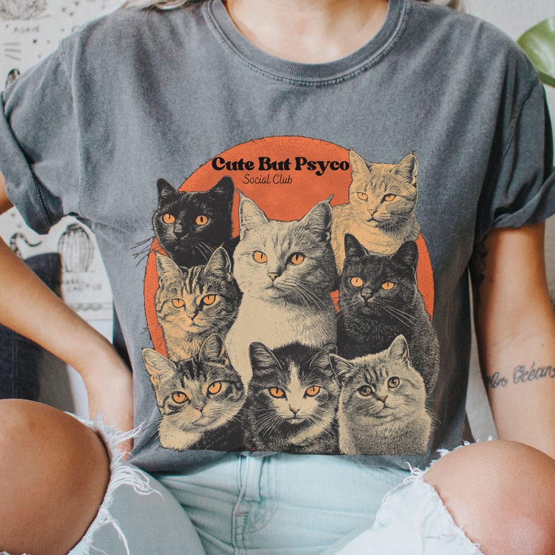 May include: A grey t-shirt with a vintage-style graphic of cats. The graphic features a group of cats with different coloured fur and the text "Cute But Psycho Social Club".