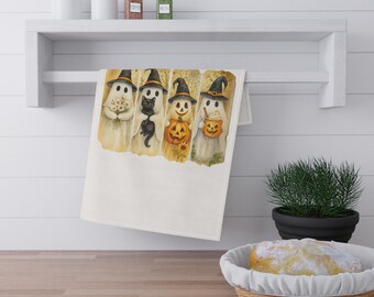 Può includere: Un canovaccio da cucina bianco con illustrazioni ad acquerello di fantasmi con cappelli da strega, ognuno con oggetti di Halloween. I fantasmi tengono fiori, un gatto nero, una zucca e un secchio di caramelle. L'asciugamano è appeso a una mensola bianca.