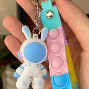 Peut inclure: Un porte-clés avec un lapin astronaute blanc avec des accents bleus et roses. Le porte-clés comprend un jouet fidget pop-it coloré avec des sections roses, bleues et jaunes. Le lapin a un écran facial et des oreilles bleues.