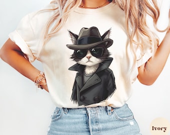 May include: Ivory-coloured t-shirt featuring a cartoon cat wearing a black fedora, sunglasses, and a black trench coat. The cat has a white face and chest. The t-shirt is paired with blue jeans. The word "Ivory" is printed at the bottom right.
