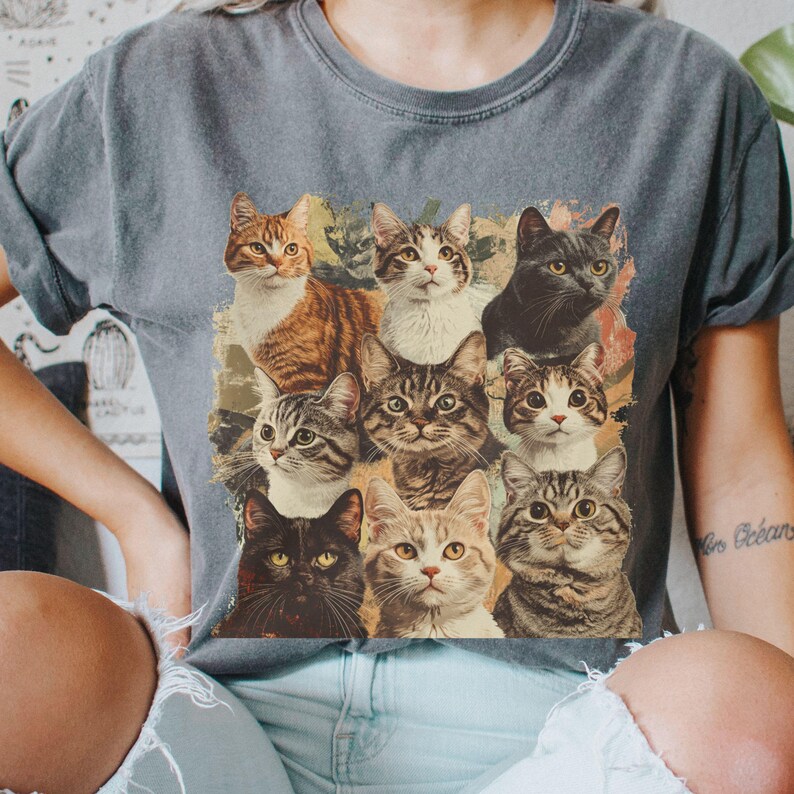 K&ouml;nnte beinhalten: Ein graues T-Shirt mit einem Vintage-Druck von neun Katzen in verschiedenen Posen. Die Katzen sind alle in verschiedenen Farben und haben unterschiedliche Gesichtsausdr&uuml;cke. Der Druck ist von einem verblassten, abgenutzten Rand umgeben.
