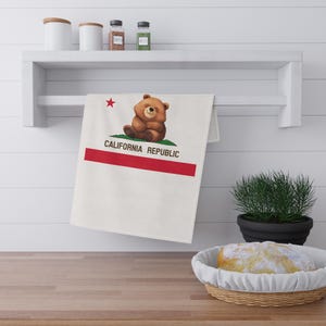 Könnte beinhalten: Weißes Küchentuch mit dem Design der kalifornischen Republikflagge. Die Flagge zeigt einen braunen Bären, einen roten Stern, einen roten Streifen und die Worte "CALIFORNIA REPUBLIC". Das Handtuch hängt an einem weißen Regal.