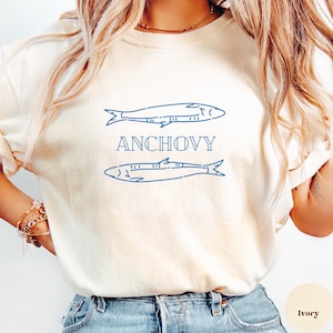 May include: Cream-coloured sweatshirt with a blue anchovy design and the word "ANCHOVY" in blue. The design features two fish illustrations. The shirt is made of a soft material and is suitable for casual wear.