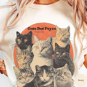 May include: A white t-shirt featuring a graphic of a group of cats in a circle with a red background. The text "Cute But Psyco Social Club" is printed on the graphic. The shirt is available in ivory.