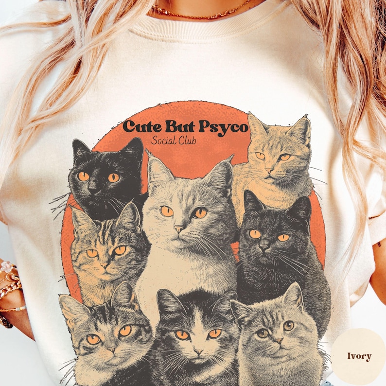 May include: A white t-shirt featuring a graphic of a group of cats in a circle with a red background. The text "Cute But Psyco Social Club" is printed on the graphic. The shirt is available in ivory.