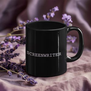 May include: A black ceramic mug with the word "SCREENWRITER" printed in white. The mug has a curved handle and is set against a backdrop of purple flowers and a lavender-colored cloth.