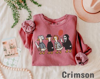 May include: Crimson-colored sweatshirt featuring a Halloween-themed graphic of four ducks dressed as horror movie characters. The ducks are holding a saw, a balloon, a knife, and wearing a Ghostface mask. The text "Crimson" is at the bottom.