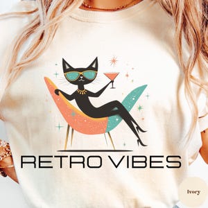Könnte beinhalten: Elfenbeinfarbenes Sweatshirt mit Retro-Design. Es zeigt eine schwarze Katze mit Sonnenbrille und Halskette, die einen Cocktail hält, während sie in einem Stuhl aus der Mitte des Jahrhunderts liegt. Der Text "RETRO VIBES" ist darunter in Schwarz gedruckt.