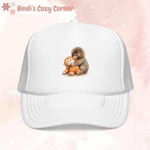 Peut inclure: Casquette de camionneur blanche avec une visière incurvée et un dos en filet. La casquette présente une illustration à l'aquarelle de deux singes qui s'embrassent. Le texte "Bindi's Cozy Corner" est imprimé au-dessus.