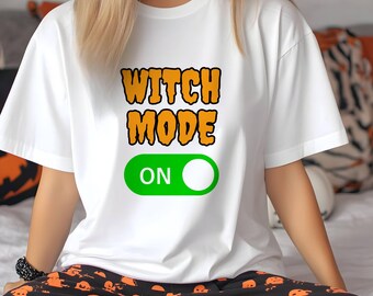 May include: White t-shirt with the text "WITCH MODE ON" in orange and black. The design includes a green on/off switch graphic. The person is wearing black pajama pants with orange Halloween-themed designs.