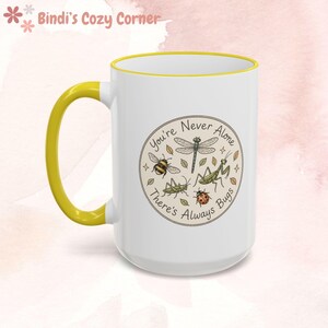 Peut inclure: Mug en céramique blanche avec une anse et un bord jaunes. Le mug présente un motif circulaire avec une libellule, une abeille, des sauterelles et une coccinelle, avec le texte "You're Never Alone, There's Always Bugs."