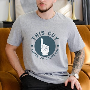 May include: A gray t-shirt with a blue circle graphic that says "This Guy Loves To Complain" with a white hand pointing up.