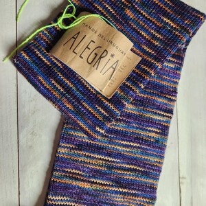 May include: A hand-knitted scarf in a vibrant mix of colors, including purple, blue, orange, and green. The scarf features the word "ALEGRIA" on a brown tag. The yarn is a blend of colors, creating a textured and visually appealing design.