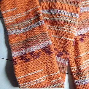 May include: An orange knitted scarf with white, purple, and brown stripes. The scarf is made of a soft, textured yarn and has a warm, autumnal color palette. The scarf is folded and draped.