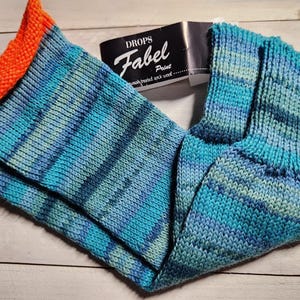 May include: A pair of knitted socks in shades of blue and teal with orange cuffs. The socks are made of Drops Fabel Print wash-treated sock wool. The socks are folded on a light wooden surface.