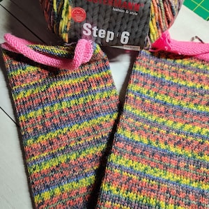 May include: A pair of knitted leg warmers in a rainbow of colors, with a bright pink cuff. A ball of multicolored yarn sits above the leg warmers. The text "Step 6" is visible on the yarn label.