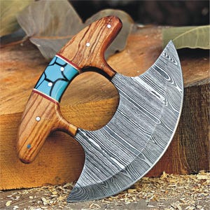 May include: A curved, stainless steel knife with a wooden handle. The handle is decorated with a blue and white inlay. The knife is a cleaver style with a Damascus pattern.