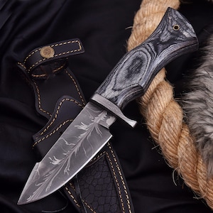May include: A handcrafted hunting knife with a patterned blade and a wooden handle. The knife is set in a black leather sheath with a decorative button. The handle has a dark gray wood grain pattern. A rope is in the background.