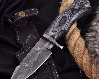 May include: A handcrafted hunting knife with a patterned blade and a wooden handle. The knife is set in a black leather sheath with a decorative button. The handle has a dark gray wood grain pattern. A rope is in the background.