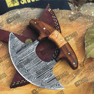 Op de afbeelding: Een Damascus stalen cleaver met een houten handvat. De cleaver heeft een gebogen lemmet en een decoratief patroon op het lemmet. De cleaver ligt op een houten oppervlak met een leren schede.