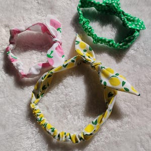 May include: Three fabric headbands with knotted bows. One headband is green with white polka dots, one is pink with white polka dots, and one is yellow with green leaves and lemons.