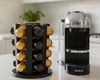 May include: A black rotating coffee pod holder filled with gold and black capsules, positioned next to a black and chrome Nespresso coffee machine with a clear water tank and the Nespresso brand name printed on the front base of the unit.