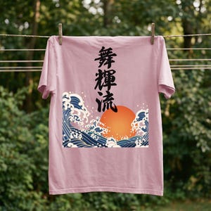 A  blossom-colored t-shirt hangs on a clothesline outdoors with wooden clothespins against a background of blurred greenery. The unique ukiyo-e-style design on the t-shirt showcases an orange sun and dynamic blue waves. Black Japanese kanji script meaning "Infinite Flow" is printed vertically.
