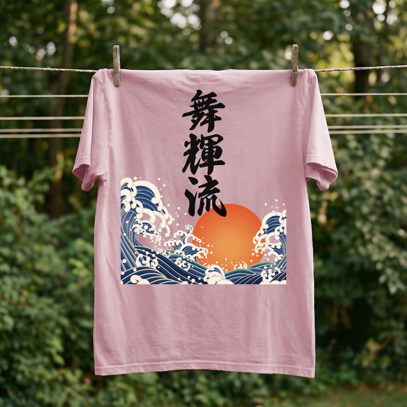 A  blossom-colored t-shirt hangs on a clothesline outdoors with wooden clothespins against a background of blurred greenery. The unique ukiyo-e-style design on the t-shirt showcases an orange sun and dynamic blue waves. Black Japanese kanji script meaning "Infinite Flow" is printed vertically.