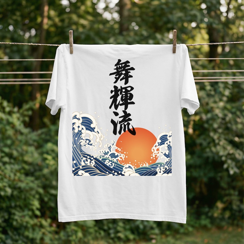 A white t-shirt hangs on a clothesline outdoors with wooden clothespins against a background of blurred trees and foliage. The t-shirt's ukiyo-e-inspired design depicts a brilliant orange sun and powerful blue waves. Black Japanese kanji script meaning "Infinite Flow" is printed vertically.
