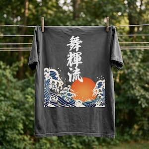 A dark gray / pepper colored, vintage-wash t-shirt hanging from a clothesline against a blurred green forest background. The shirt features a large print on the front with vertical white Japanese Kanji characters positioned above a traditional Japanese art graphic depicting a rising orange sun and crashing blue and white ocean waves.