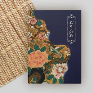May include: A notebook with a Japanese-inspired design. The cover features a floral pattern with pink and white blossoms, a green bird, and the Japanese text "おりびわ" in a white decorative frame against a navy blue background.