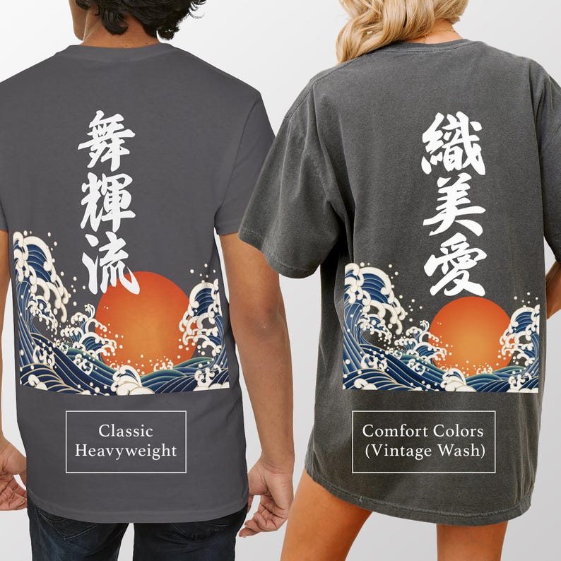 Back view of two people modeling dark gray t-shirts to compare fabric styles. The person on the left wears a smooth "Classic Heavyweight" t-shirt with white vertical Kanji and a wave-and-sun design. The person on the right wears a textured "Comfort Colors (Vintage Wash)" t-shirt with different white vertical Kanji and the same wave-and-sun design.