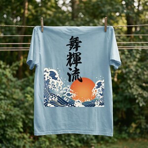 A light blue t-shirt hangs on a clothesline outdoors with wooden clothespins. A forest is blurred in the background. The t-shirt's dynamic ukiyo-e style design depicts an orange sun and dramatic blue waves. Black Japanese kanji script meaning "Infinite Flow" is printed vertically.