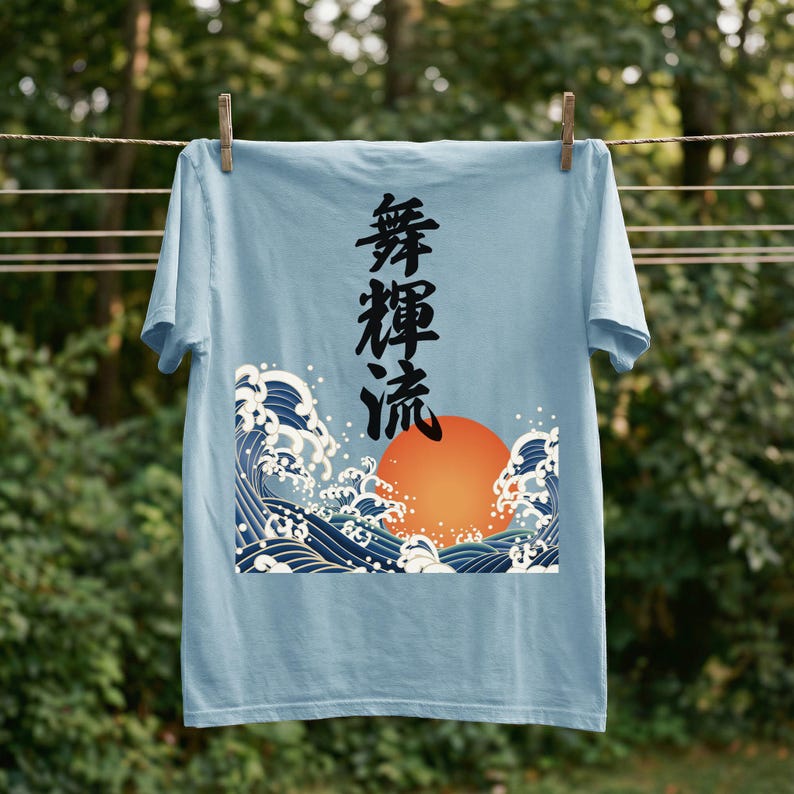A light blue t-shirt hangs on a clothesline outdoors with wooden clothespins. A forest is blurred in the background. The t-shirt's dynamic ukiyo-e style design depicts an orange sun and dramatic blue waves. Black Japanese kanji script meaning "Infinite Flow" is printed vertically.